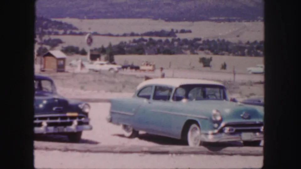 A 1958 Colorado home-movie frame shows classic cars at a rural roadside stop with open fields and small structures behind them.