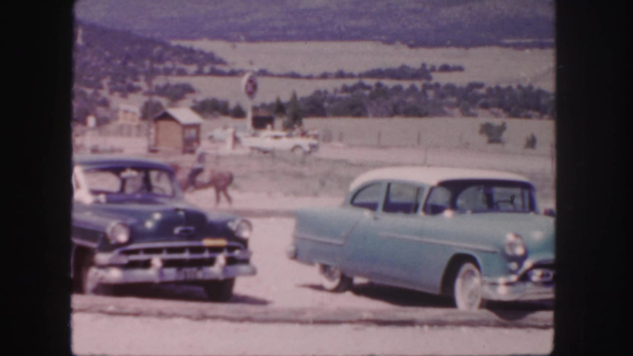 Two vintage cars pass a horseback rider near an open hillside.