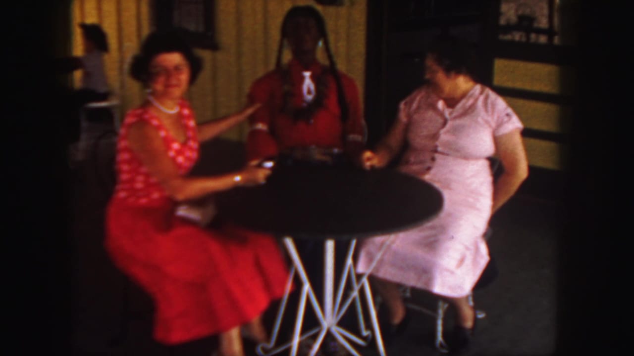 In a 1958 Colorado 8mm home-movie frame, two women in red and pink dresses sit beside a braided life-size doll in red.