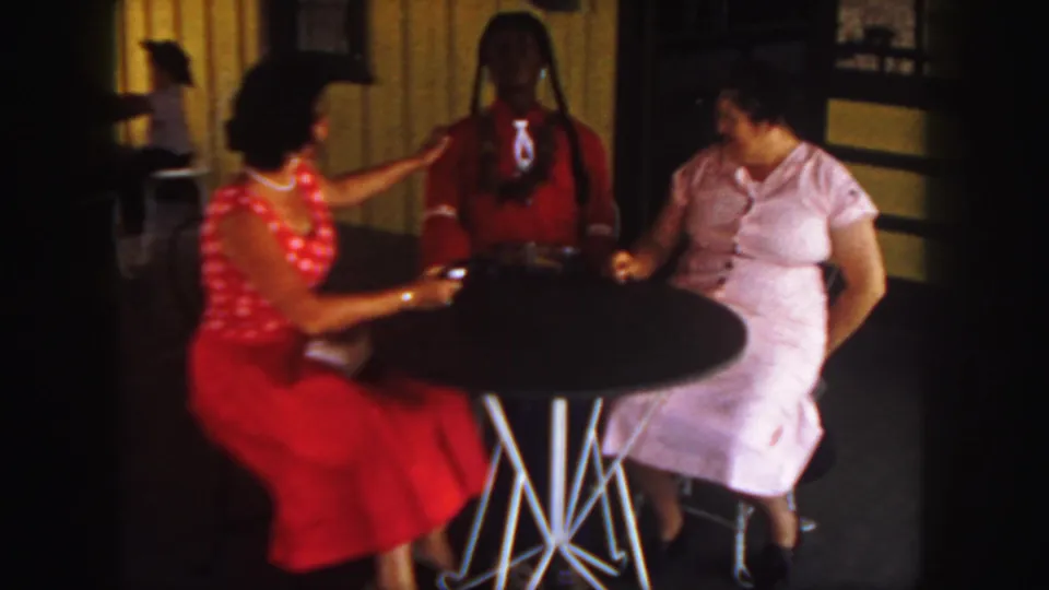 The frame shows the woman in red reaching toward the braided doll as the woman in pink looks on in the dim interior.