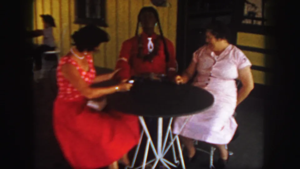 In the home-movie interior, both women face the table while the red-clothed doll stands upright between their chairs.