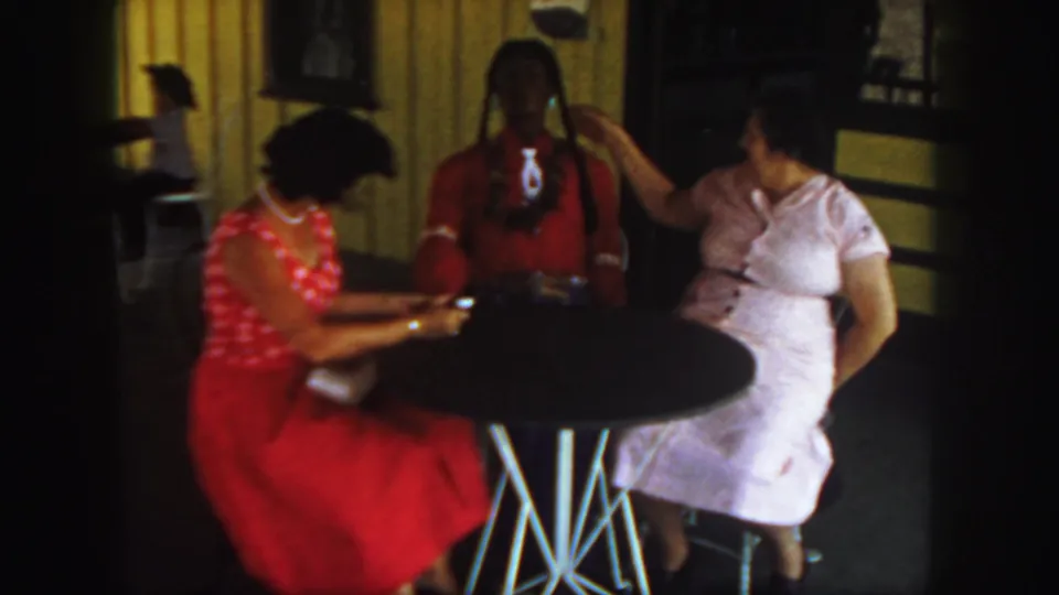 The woman in pink lifts a hand toward the braided doll as the woman in red looks down near the table in the 1958 Colorado scene.