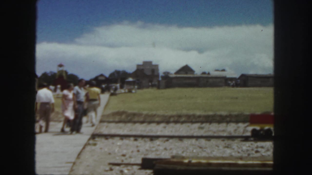 1958 Colorado 8mm frame showing adults on a walkway near a miniature train track with rustic buildings in the distance.
