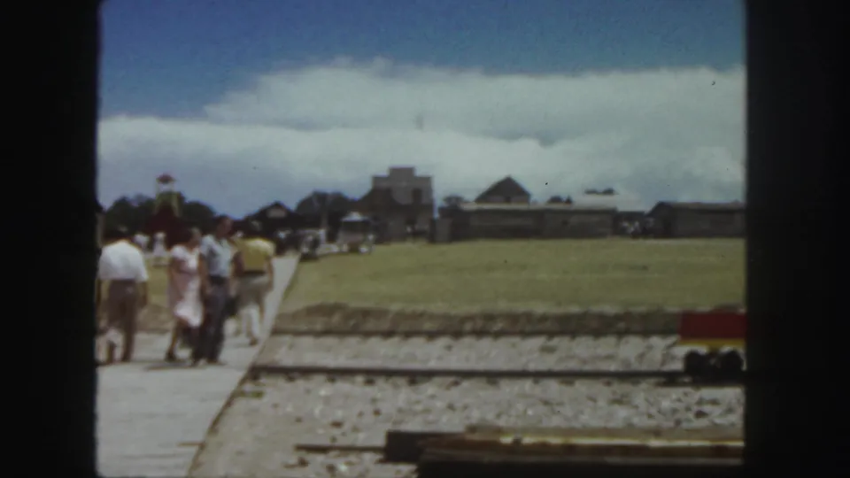 1958 Colorado 8mm frame showing adults on a walkway near a miniature train track with rustic buildings in the distance.
