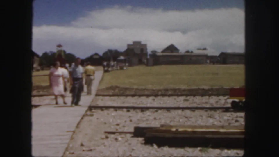 1958 home-movie frame of visitors walking near a miniature railway, with farm-like buildings across an open Colorado field.