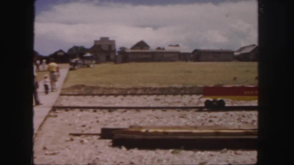 A 1958 Colorado 8mm frame shows visitors receding along a path near miniature tracks, open grass, and distant rustic structures.
