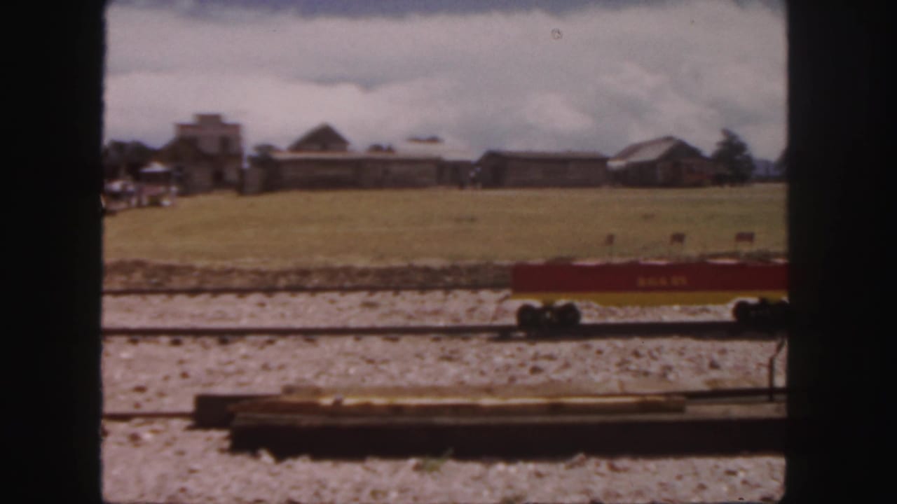 Small red Wells Fargo rail car on gravel track with open field and rustic buildings behind it.