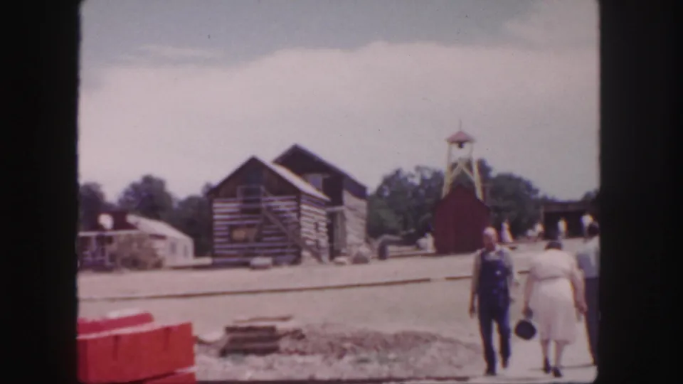 1958 Colorado 8mm frame with visitors walking near log-style cabins, a small tower, track area, and red barriers at an outdoor site.