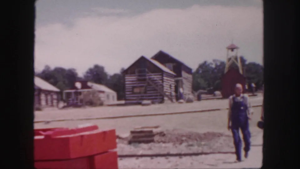 1958 Colorado home-movie frame showing a man walking past log cabins, red barriers, a gravel track area, and a small tower.