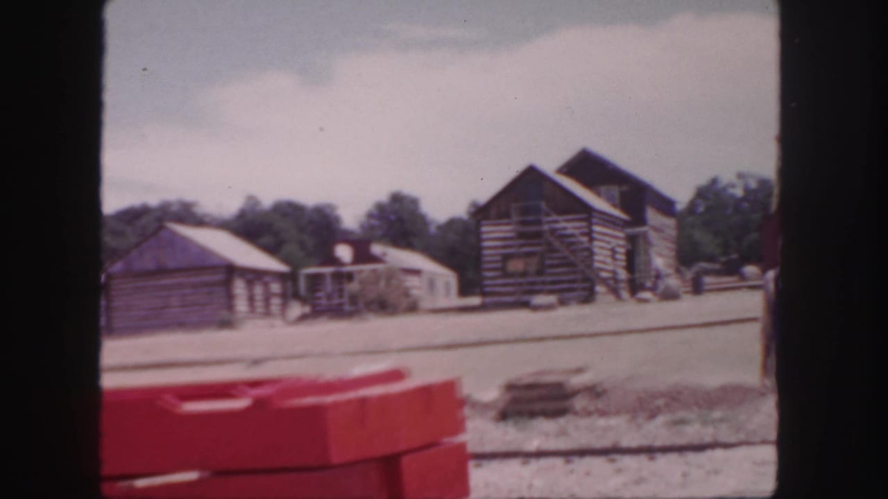 1958 Colorado 8mm frame showing rustic log buildings, trees, dusty ground, and a red foreground barrier or object.