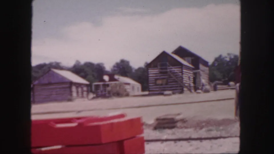 1958 Colorado 8mm frame showing rustic log buildings, trees, dusty ground, and a red foreground barrier or object.