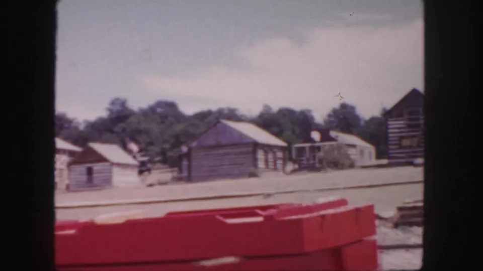 1958 Colorado home-movie view of weathered wooden cabins, trees, and a red object across a sunlit dusty area.