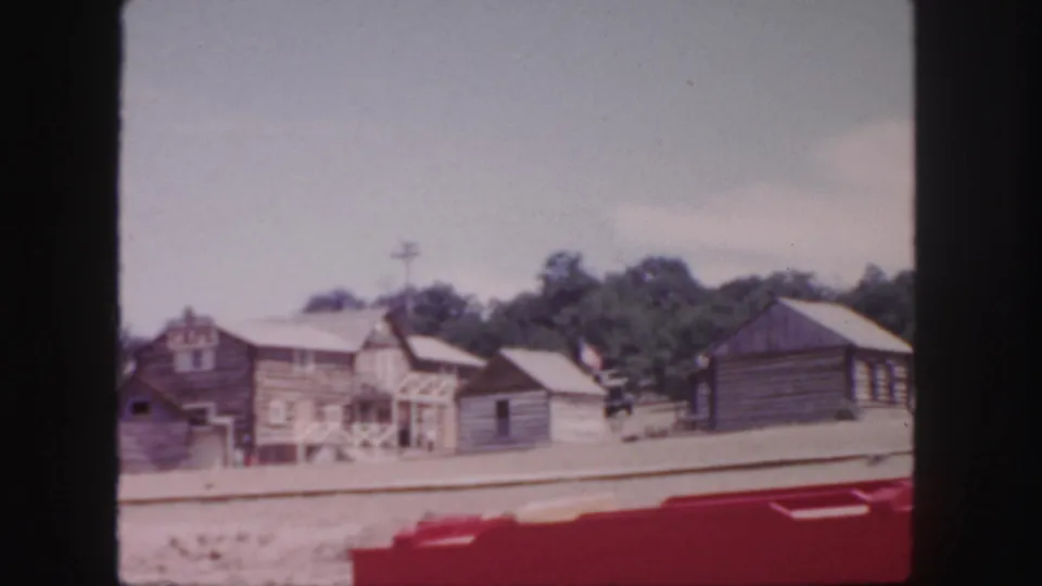 1958 Colorado frame with multiple rustic wooden buildings, a small shed, dusty ground, and trees in the background.