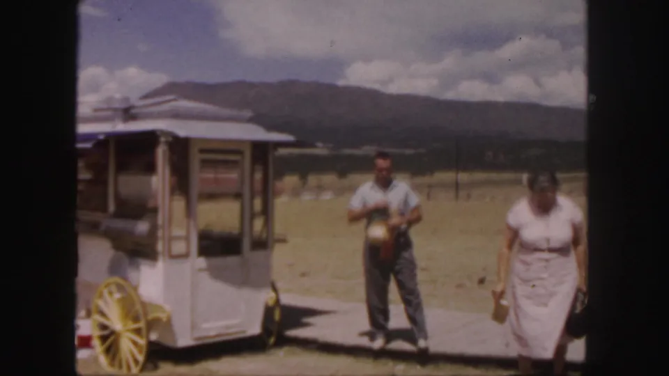 1958 Colorado 8mm frame showing two adults near a white vending-style cart with yellow wheels and distant mountains.