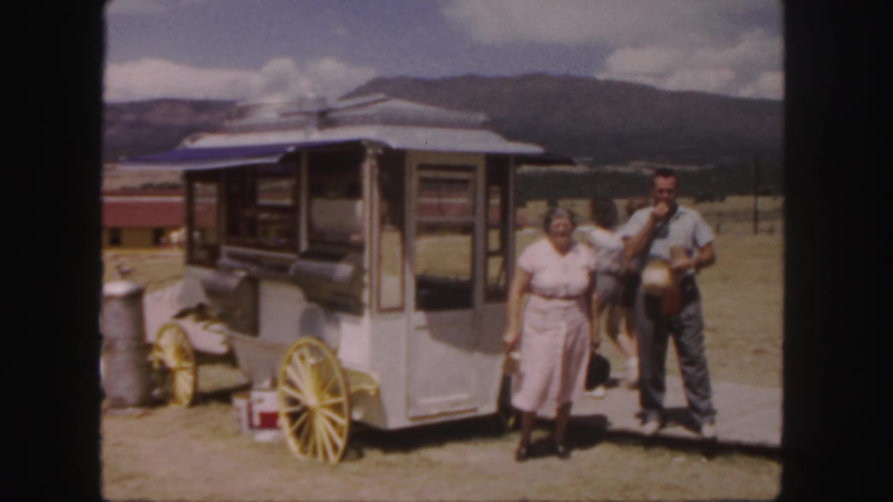 Woman and man stand beside a wheeled food stand with mountains behind them.