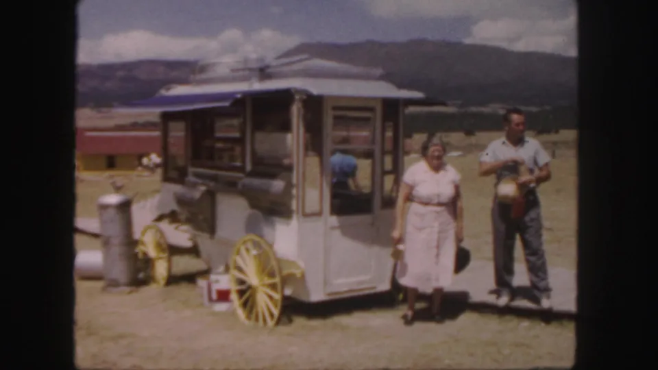 A woman and man pause beside a small wheeled food stand in Colorado, with yellow cart wheels, a blue awning, and hills in the background.