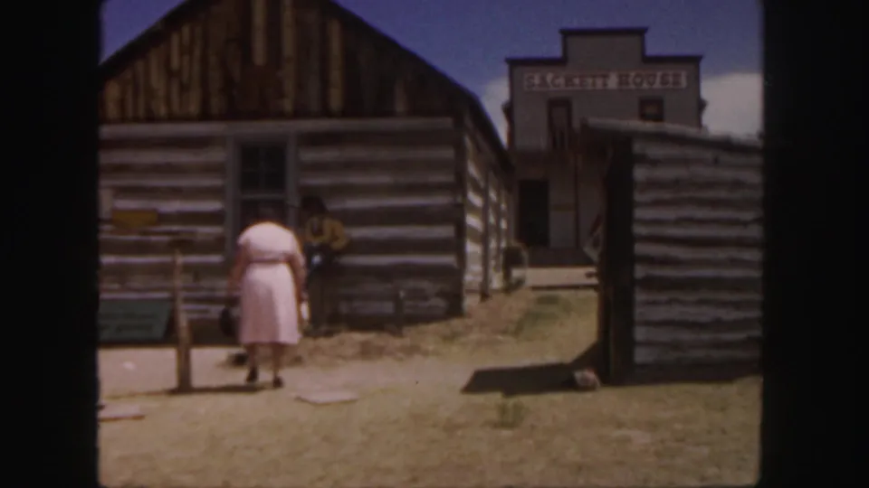 The scene shifts to a rustic western-style street where a woman in pink walks near log cabins and a building marked Sackett House.