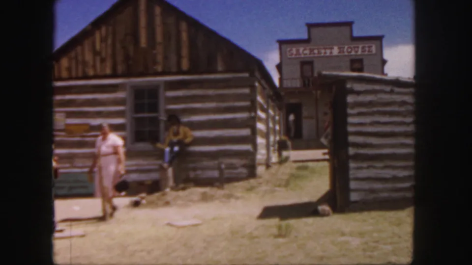 A woman walks through the Colorado western-town setting while a man in period-style clothing stands near a log cabin and Sackett House sign.