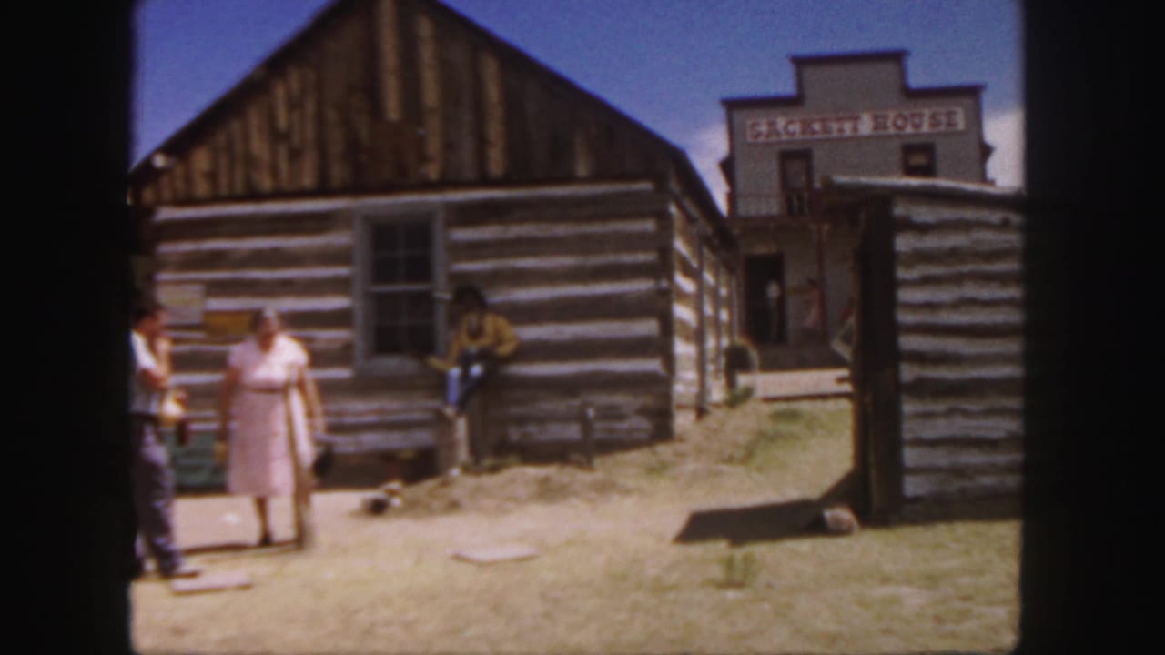 Adults stand outside a log cabin with Western village buildings in the background.