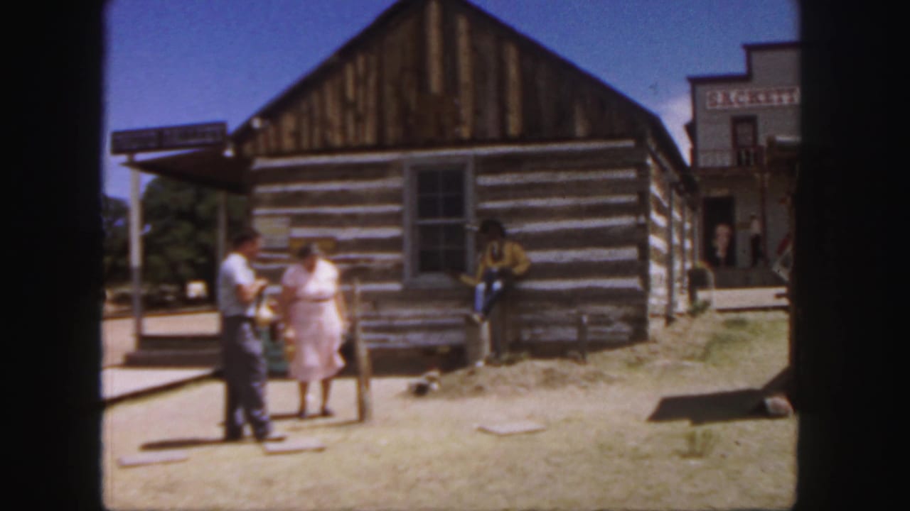 Adults talk near a log cabin while a cowboy-dressed man leans by the wall.