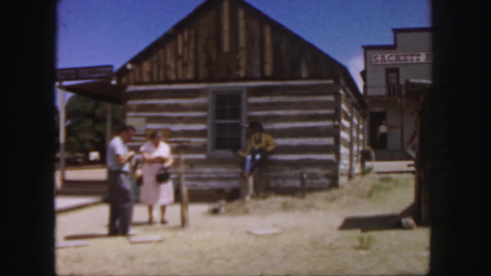A 1958 Colorado home-movie view of adults beside a weathered log cabin, with Western-themed storefronts in the background.
