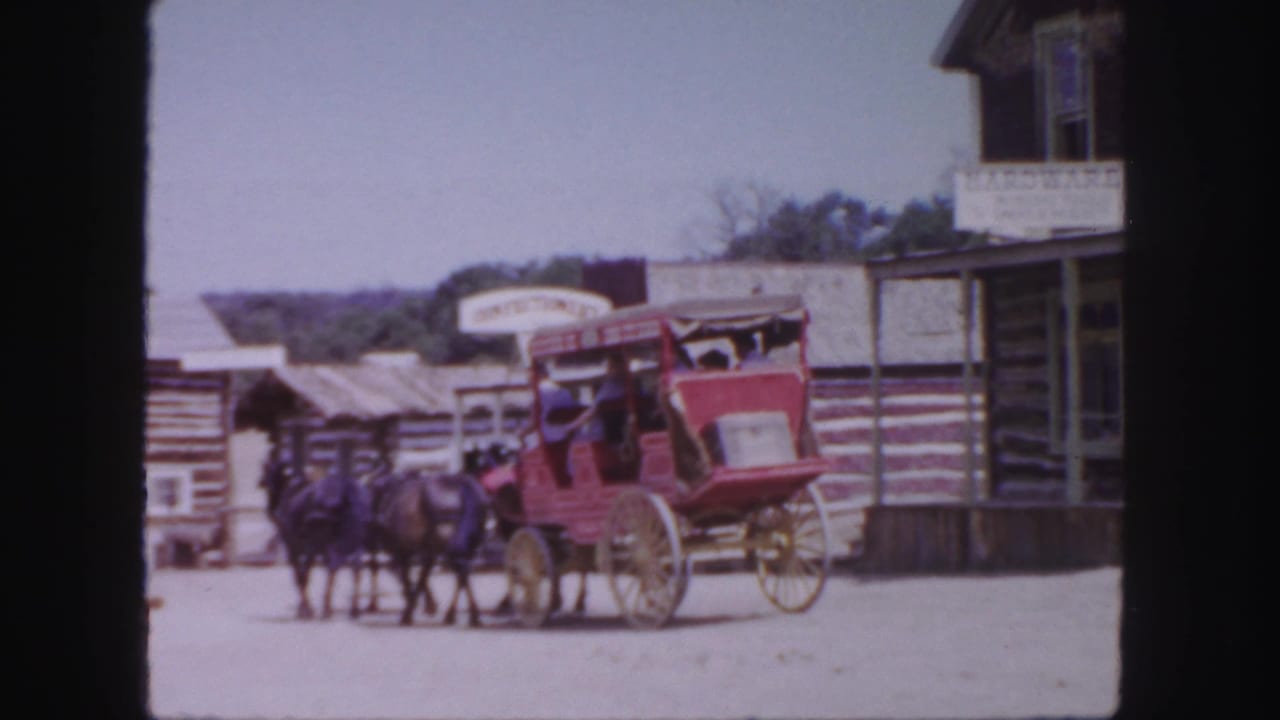 Red horse-drawn stagecoach passing log-style storefronts on a dusty Western street.