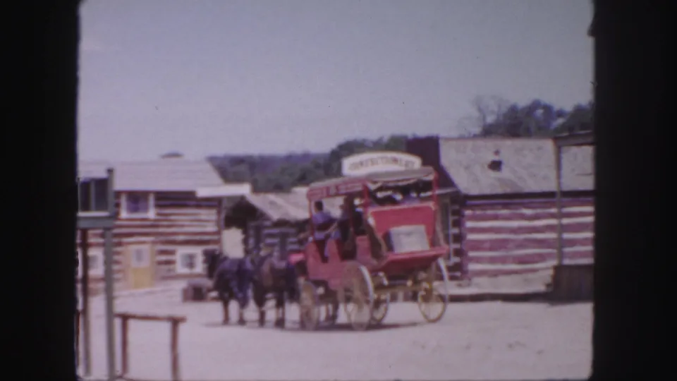 The red horse-drawn coach continues along a dusty Colorado Western town street, with passengers and painted storefront signs visible.
