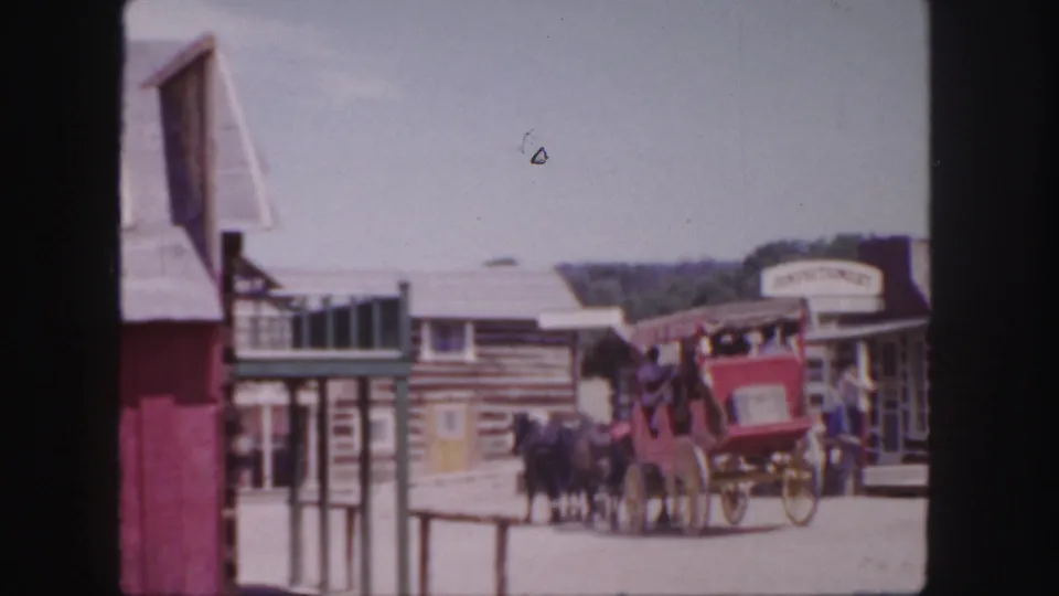A visitor-shot 8mm view shows the stagecoach moving away through the Western set, with railings and a building edge in the foreground.