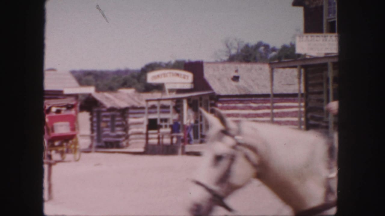 In 1958 Colorado home-movie footage, a horse and rider move past log-cabin storefronts with a confectionery sign and a red carriage nearby.
