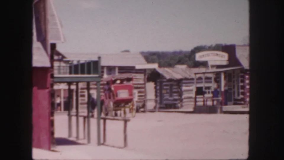 A Colorado Western-themed town scene shows log-cabin storefronts, a red carriage, pedestrians, and visible shop signs in 1958 8mm footage.