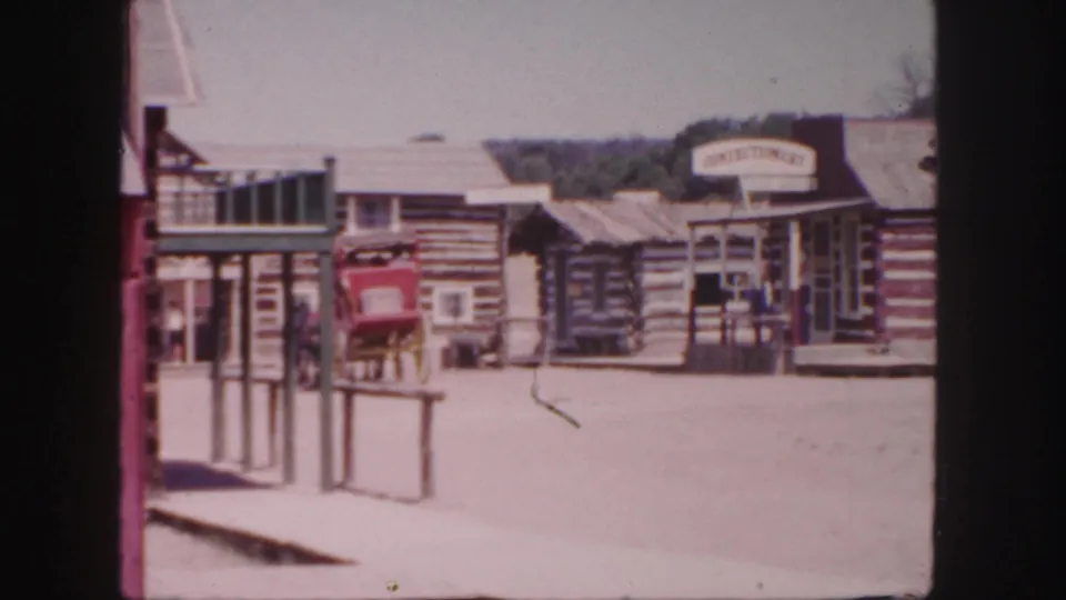 The 1958 Colorado frame shows visitors near a red carriage, log storefronts, porch posts, and a hardware-style sign along a dusty street.