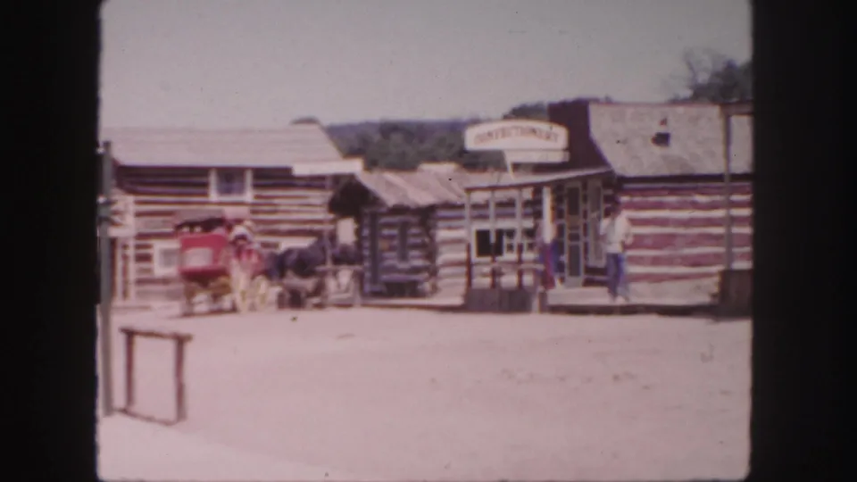 A horse-drawn red carriage, log storefronts, shop signage, and a man on the porch appear in this 1958 Colorado Western attraction scene.
