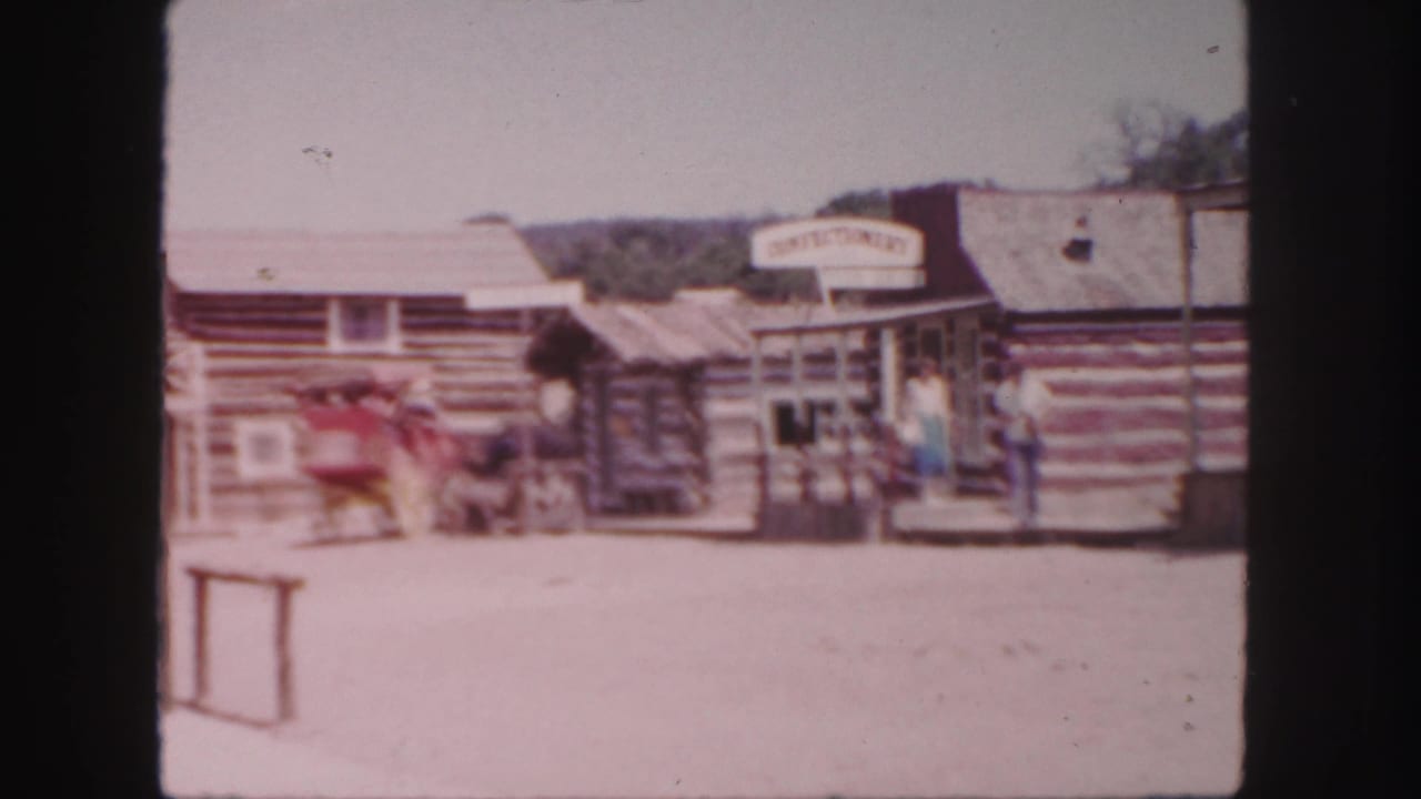 1958 Colorado home-movie frame showing Western-style log buildings, a confectionery sign, visitors, and a red wagon on a dusty street.