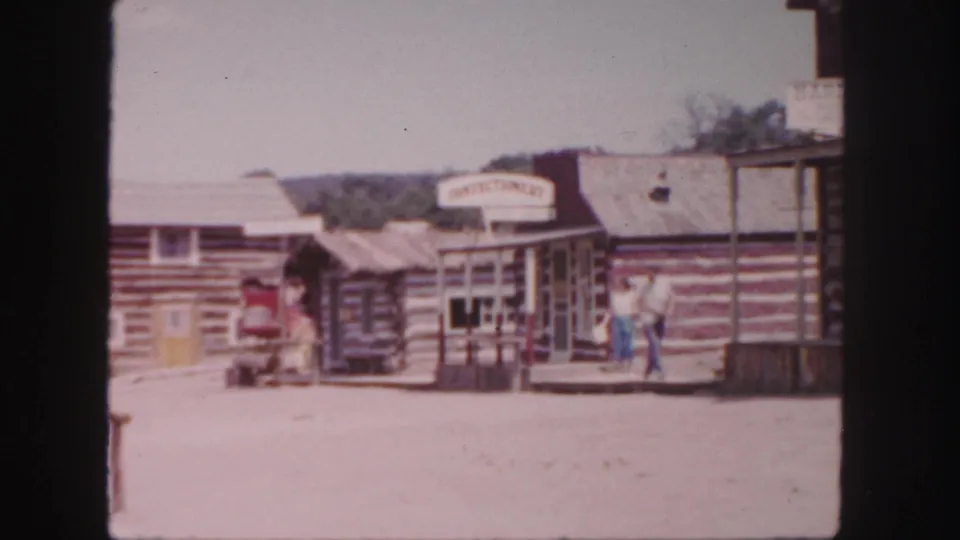 1958 Colorado frame of a Western-style street with log facades, a confectionery sign, visitors, and a red wagon in bright sun.