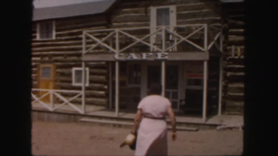 1958 Colorado home-movie view of a woman approaching a Western-style log cafe with white porch railings and a shaded entrance.