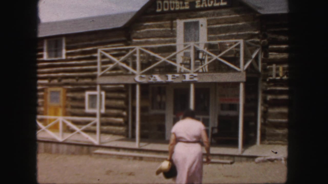 In 1958 Colorado footage, a woman in a pink dress approaches a rustic log cabin cafe with white railings and visible signage.
