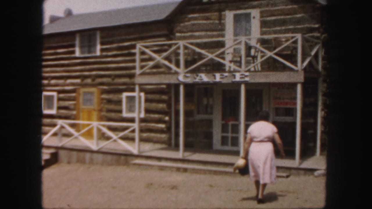 Woman nears the steps of a log cabin cafe with white railings and signs.