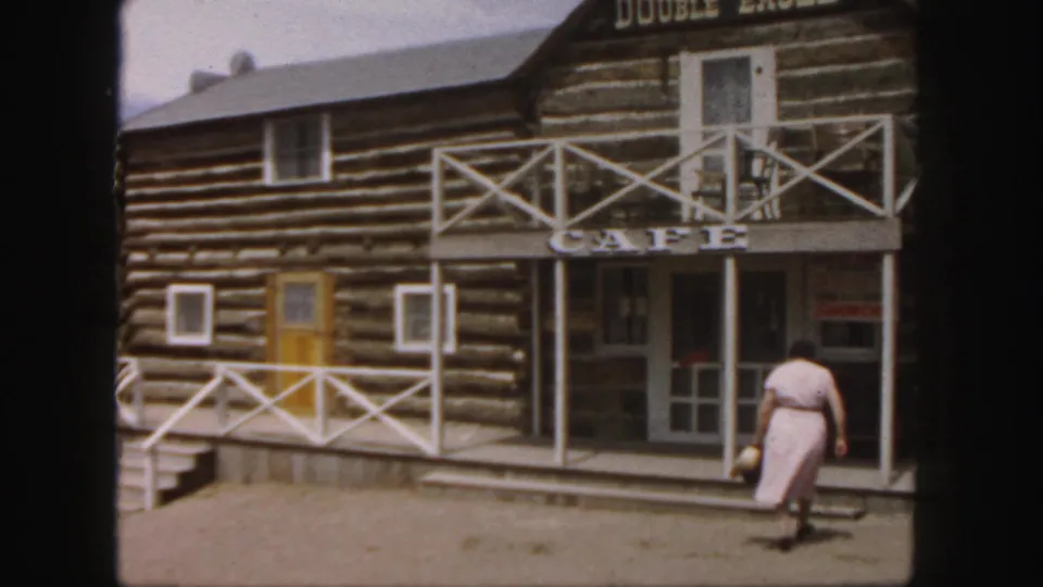 A woman in a pink dress reaches the steps of the Double Eagle cafe, framed by log walls, white railings, and an open entrance.