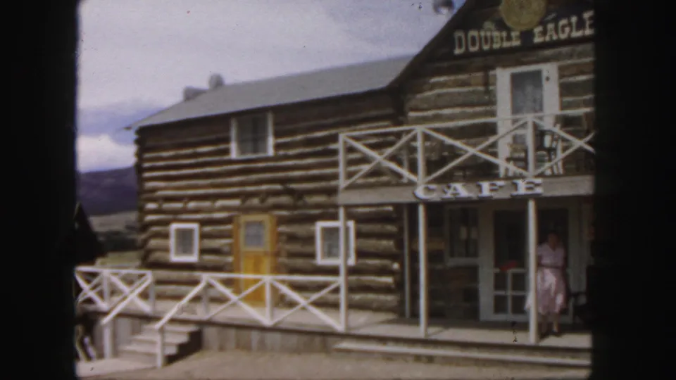 1958 Colorado frame of the Double Eagle Cafe porch, with a woman in a pink dress near the entrance and log walls behind her.