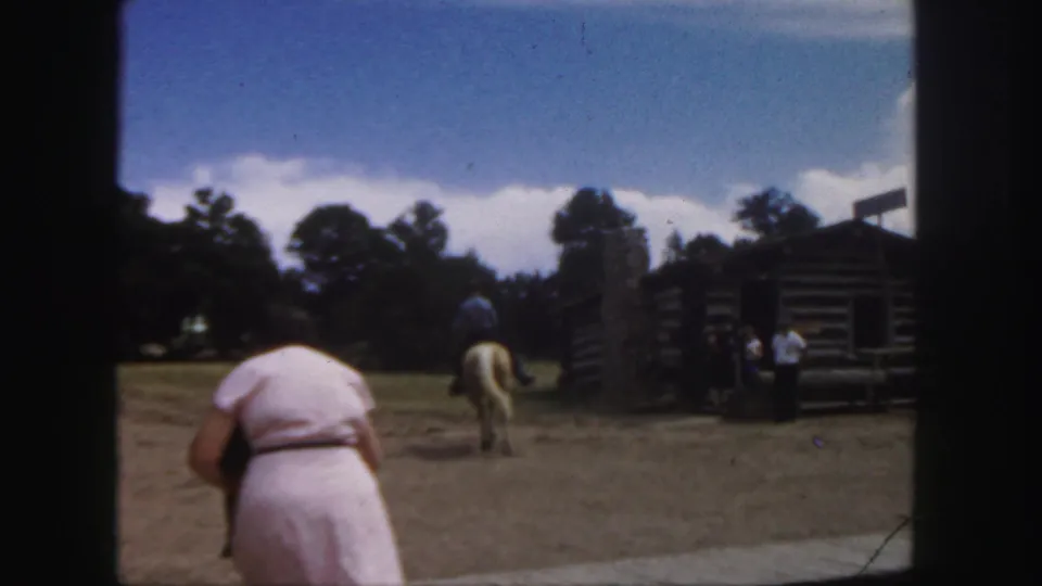 1958 Colorado home-movie frame with a woman in pink in the foreground and a horseback rider crossing an open yard near log buildings.