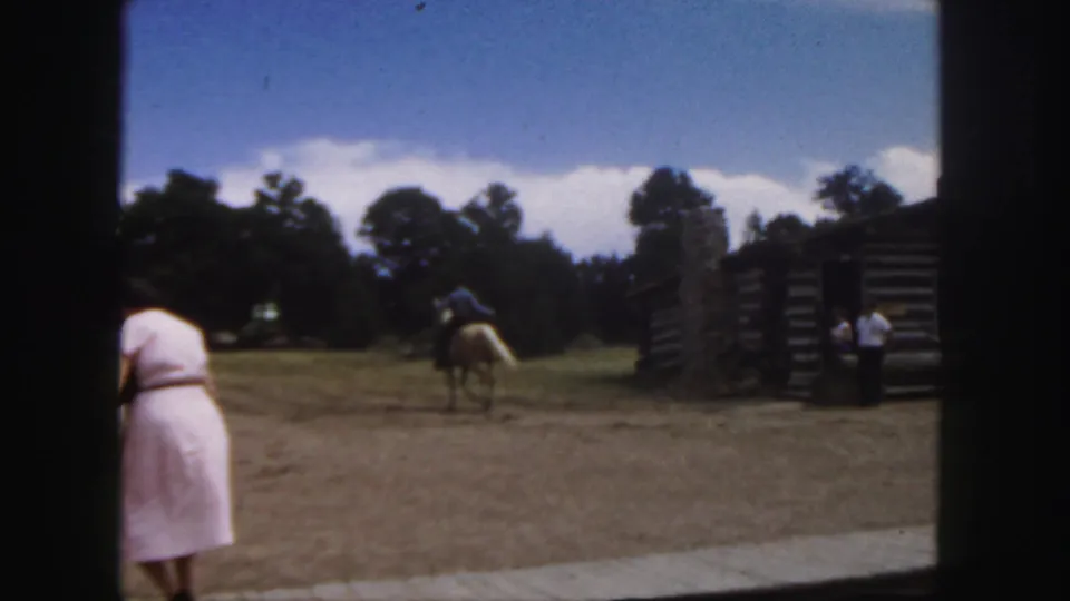 1958 Colorado frame showing a horseback rider moving away across a dusty yard beside rustic log structures and trees.