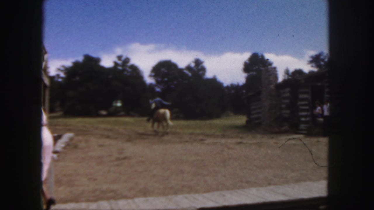 1958 Colorado home-movie frame showing a rider in a blue shirt on a tan horse moving across a dusty clearing near rustic log buildings.