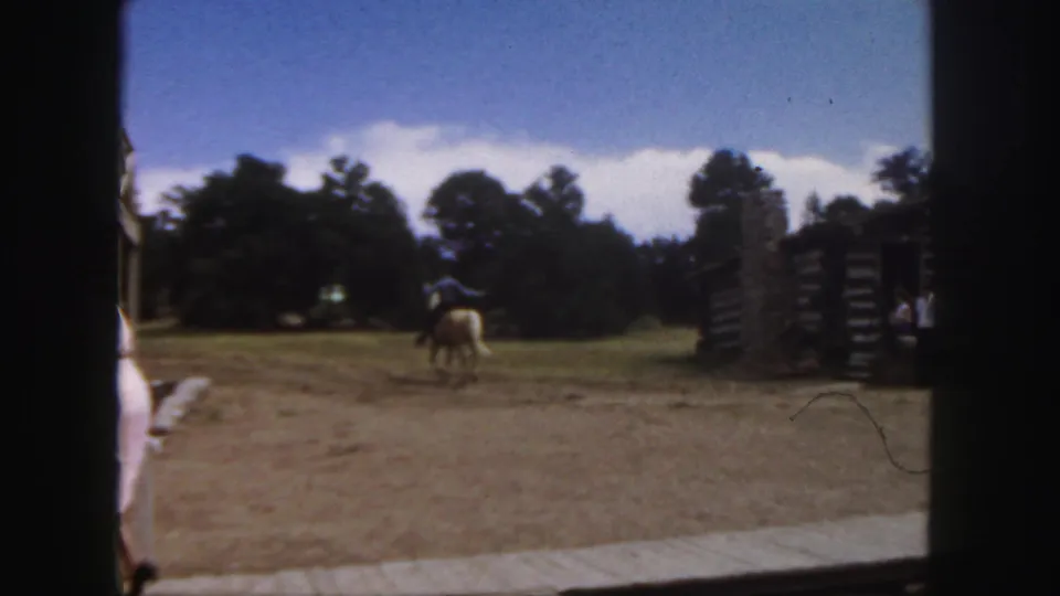 1958 Colorado home-movie frame showing a rider in a blue shirt on a tan horse moving across a dusty clearing near rustic log buildings.