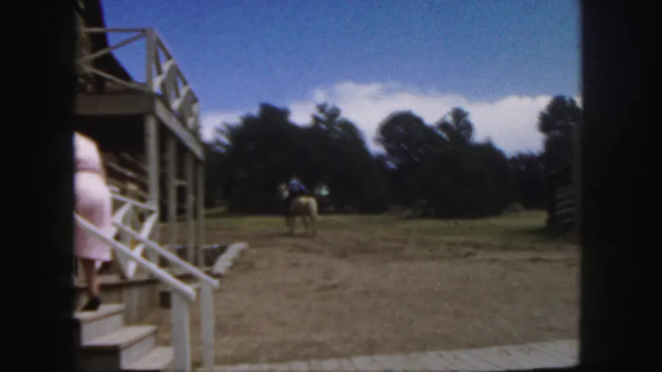 1958 Colorado frame with a porch, white railing, and a rider on a tan horse crossing the open ground near trees and log structures.
