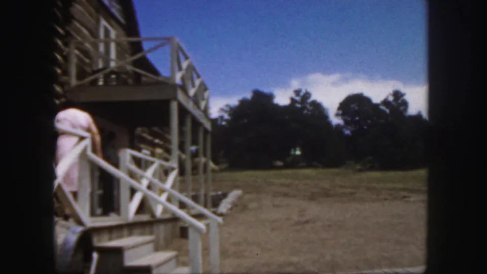 1958 Colorado home-movie frame showing a woman in a pink dress near a two-story porch overlooking a rustic settlement clearing.