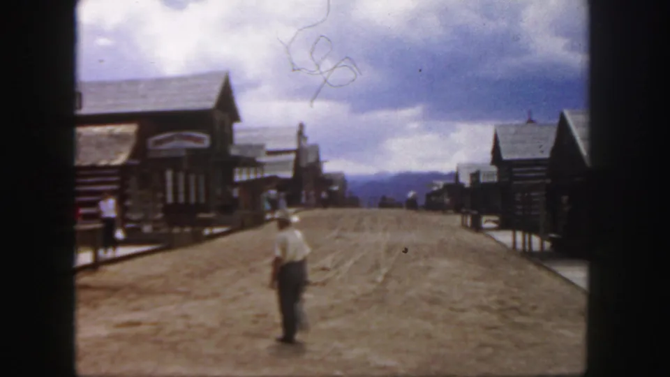 1958 Colorado frame showing a person walking along a broad dusty road through a Western-style settlement with log buildings and distant hills.