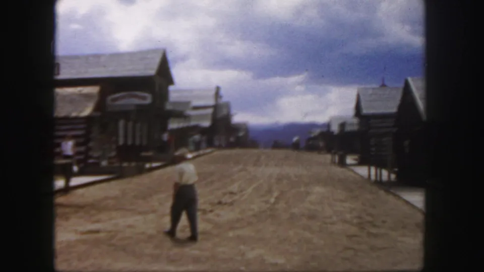 1958 Colorado home-movie frame showing a man in a light shirt and cowboy hat walking along an Old West-style dirt street.