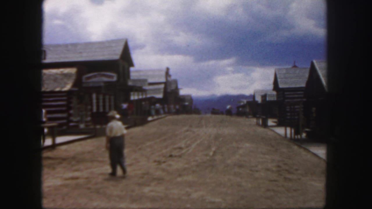 Man in a cowboy hat stands or walks near the center of a dirt street with wooden buildings nearby.