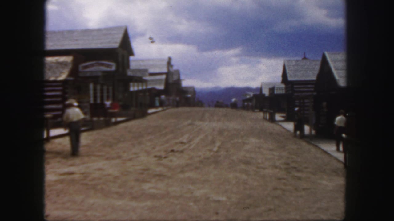 1958 Colorado 8mm frame showing a broad unpaved Western-style town street with log-front buildings, cloudy sky, and visitors along the sidewalks.