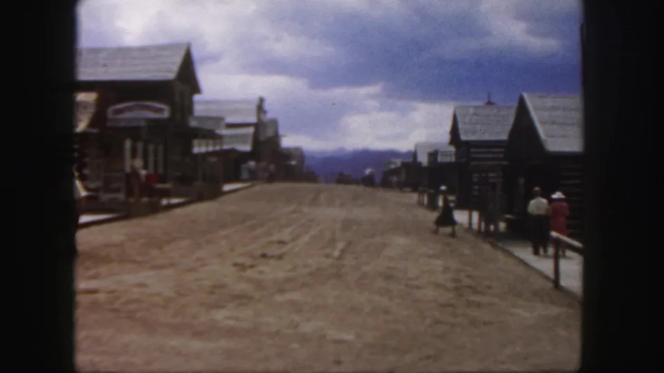 1958 Colorado home-movie frame of people strolling through a Western-style town street with rustic buildings and mountains or hills in the distance.